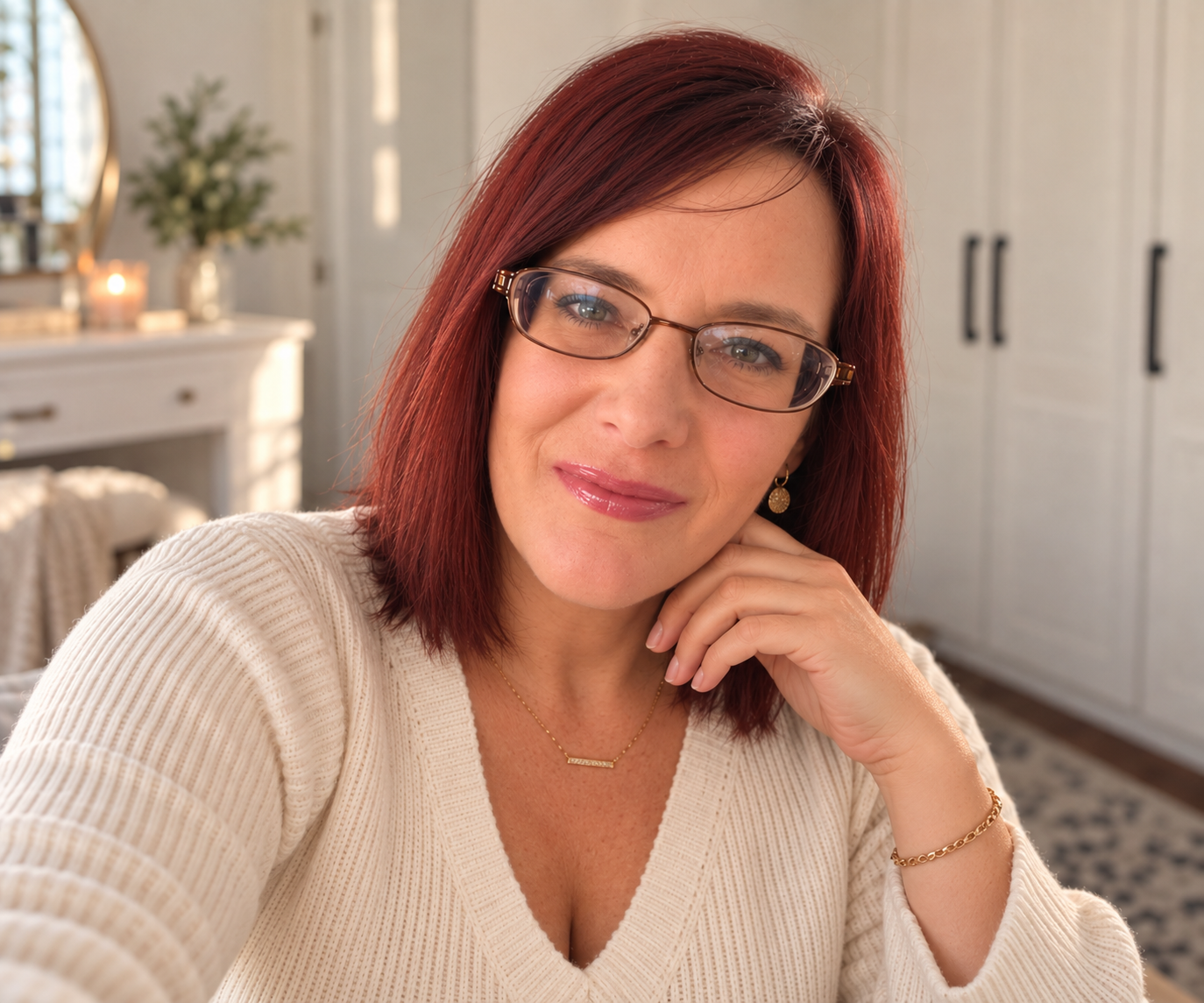 A woman with shoulder-length auburn hair and glasses smiling with her hand resting on her chin.